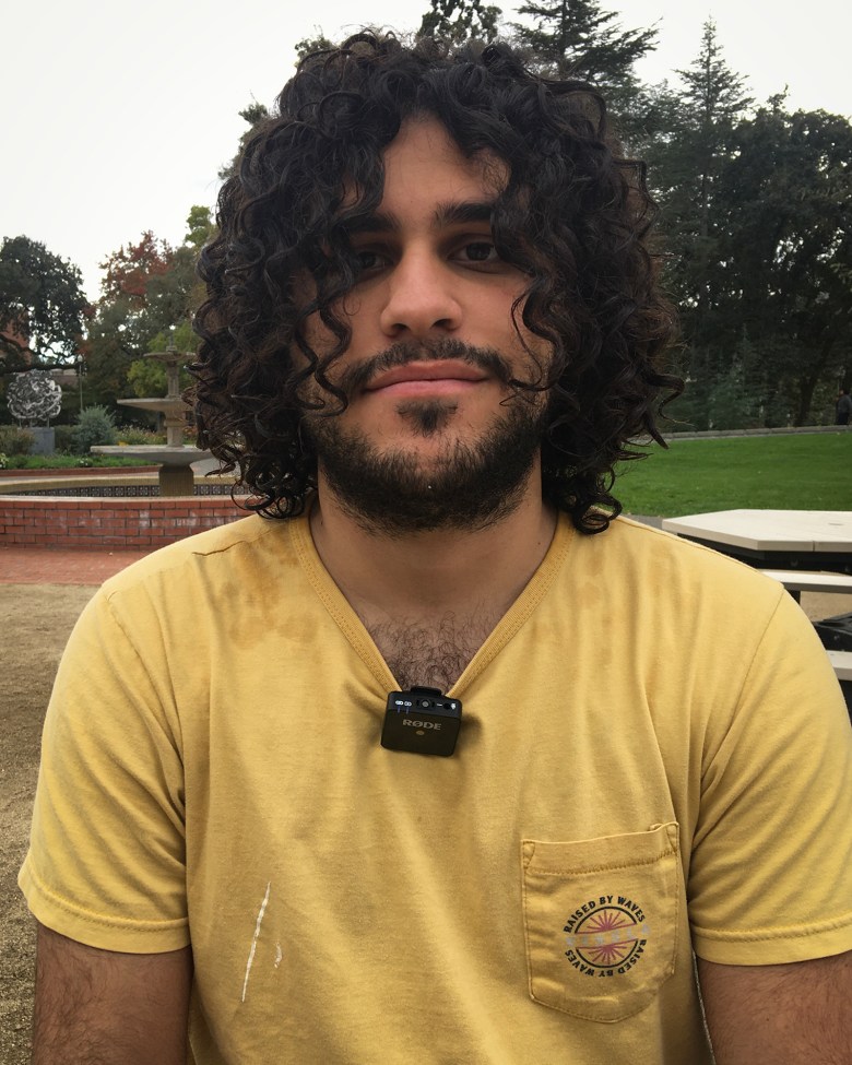 A person with chin length curly hair, a mustache and beard, stares intently into the camera. They are wearing a yellow t-shirt, and on their collar is a small, squared, microphone with the words "RODE" printed on it.