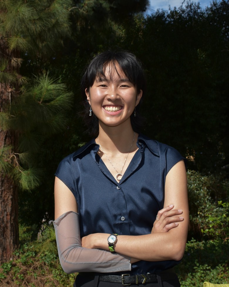 A person in a blue button up shirt, with the top button undone, crosses their arms across their body, as they smile into the camera. Behind them, a tree and verdant shrubbery is visible.