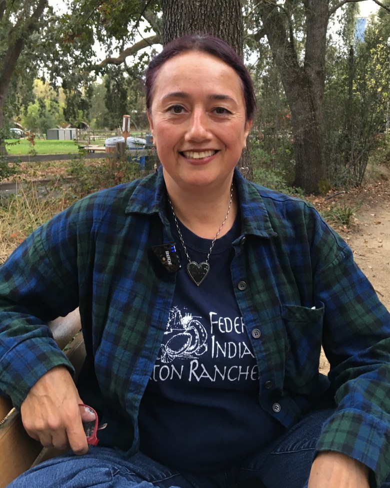 A person sits outdoors, smiling warmly at the camera. They are dressed in a dark blue T-shirt under a green and blue plaid flannel shirt. A heart-shaped pendant hangs around their neck, and they hold a pair of glasses in one hand. The background reveals trees and a fenced area.