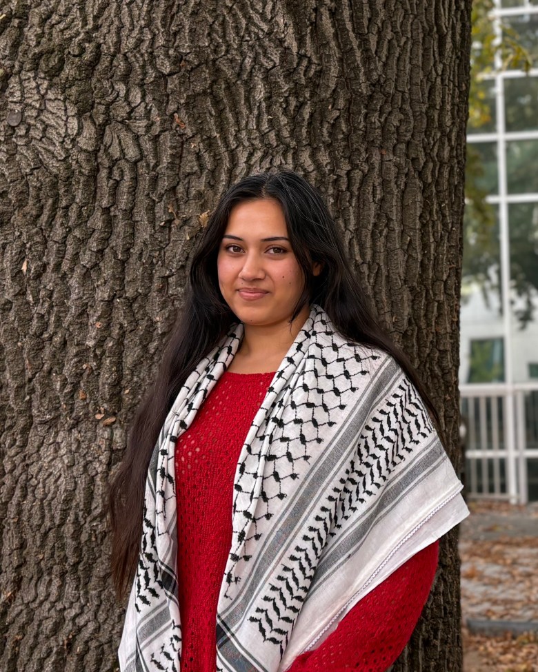 A person stands outdoors in front of a large tree trunk, wearing a red sweater and a keffiyeh scarf with a black and white pattern. They have long dark hair, and their expression is calm and warm. The background shows blurred foliage and a building with windows, suggesting an autumn setting.