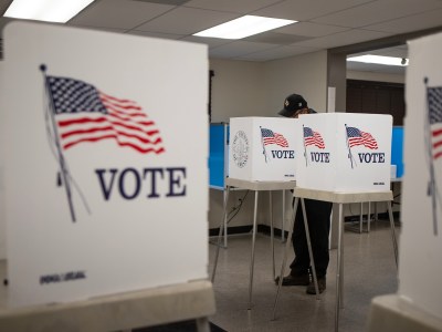 Four white voting booths with American flags and the word "VOTE" emblazoned on the side are visible. A person wearing a baseball cap casts a ballot in one of the booths. They're located in a room, reminiscent of a community center.