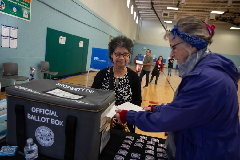 A person with short, curly black hair and wearing a black cardigan with a white and black patterned short watches as an election worker wearing a purple sweatshirt inserts a ballot in a ballot box at a vote center inside a gymnasium.