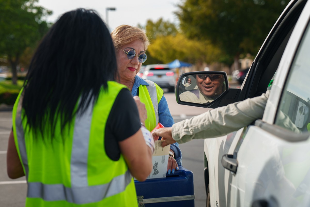 A person driving a car extends their hand out the window to insert their ballot envelope into a drop box being held by an election worker. The back of another election worker can be seen nearby.