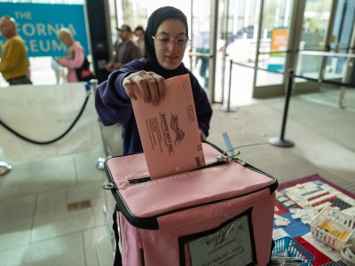 A person wearing a hijab and purple sweatshirt inserts a pink ballot into a pink ballot box at a polling place.