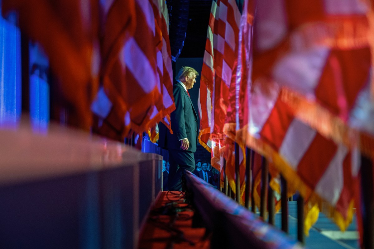 A side profile of a person in a suit and red tie walking amidst large, partially blurred flags in a dimly lit setting. The scene has a dramatic, almost solemn ambiance, with vivid red and blue lighting casting reflections on the surrounding flags, creating a sense of depth and movement.