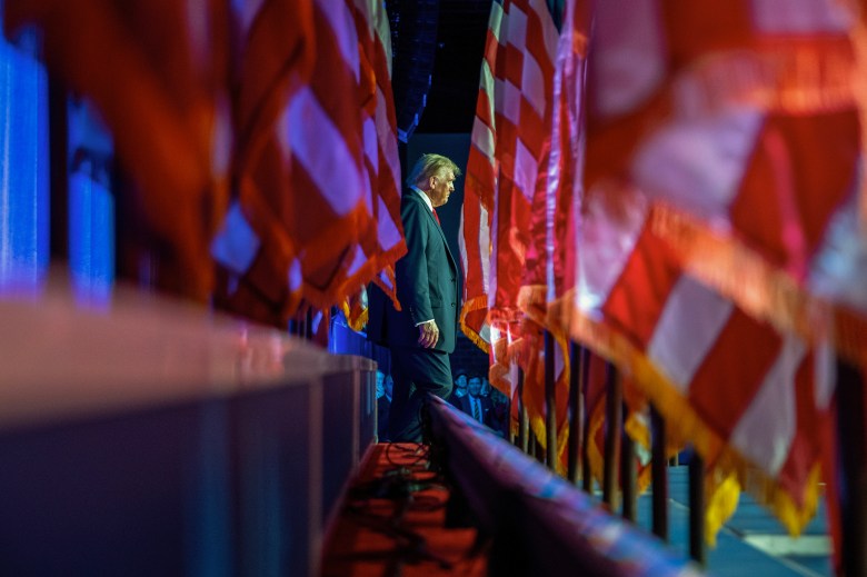 A side profile of a person in a suit and red tie walking amidst large, partially blurred flags in a dimly lit setting. The scene has a dramatic, almost solemn ambiance, with vivid red and blue lighting casting reflections on the surrounding flags, creating a sense of depth and movement.