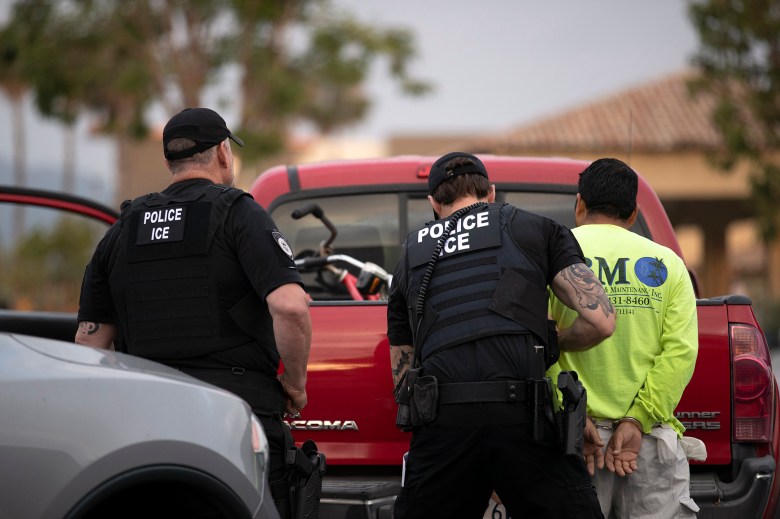 An officer searches a man in a yellow shirt handcuffed and leaning against a red truck, as another officer watches them.