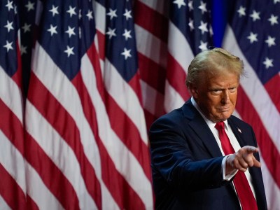 A person wearing a dark suit and red tie stands in front of a background filled with American flags. The individual is pointing forward with a determined expression, emphasizing a stance of confidence and focus amidst the patriotic setting.