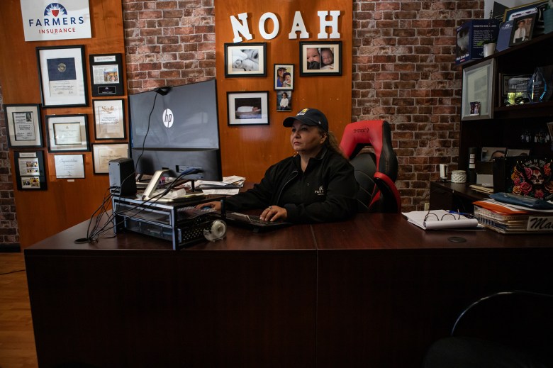 A person wearing a black hate, sweatshirt and shirt sits in front of a desk looking at a computer monitor at an insurance office. Family photos and certificates are hung up in the brick wall behind them.