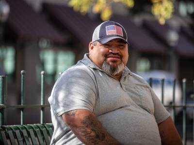 A person wearing a hat with an American flag and a grey shirt sits on a green bench in an outside area of downtown Merced.