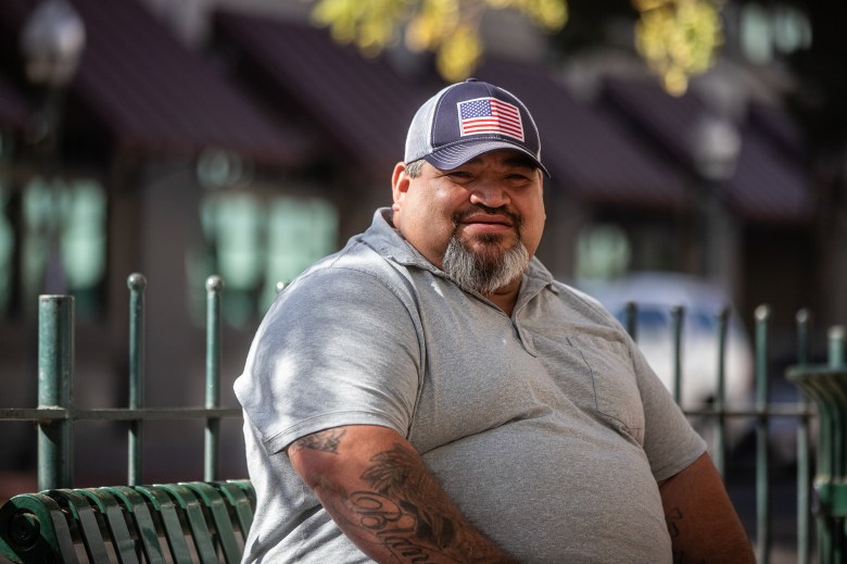 A person wearing a hat with an American flag and a grey shirt sits on a green bench in an outside area of downtown Merced.