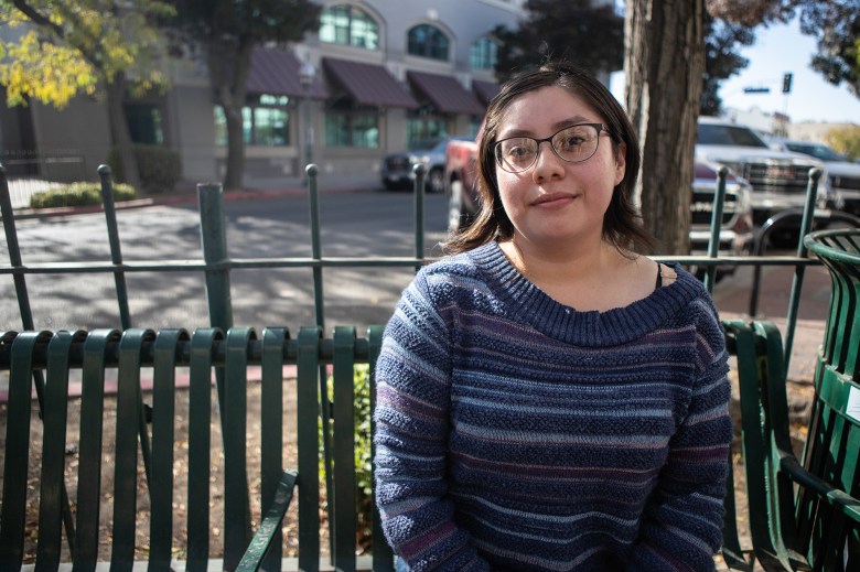 A close up view of a person wearing a blue shirt sits on a green bench outside a downtown area in Merced.