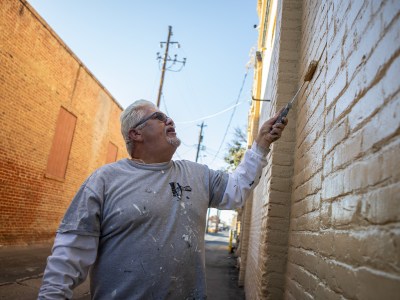 A person with white hair and wearing a grey shirt uses a paint roller to paint a yellow brick wall in an alley.