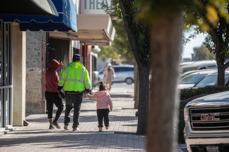 Two people walk next to each other while one is holding the hand of a child with a pink sweatshirt as they walk down a cobblestoned street next to several storefront businesses in a downtown area during the day.