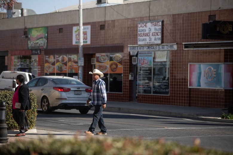 A wide view of a person wearing a white cowbiy hat, blue flannel shirt and black pants crossing an intersection in a downtown area in Delano.