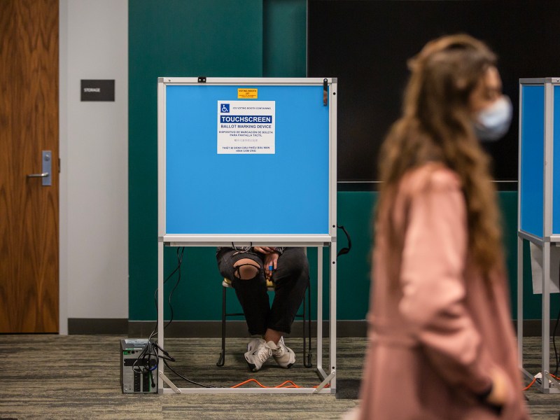 A person is seated in a wheelchair-accessible voting booth with a blue privacy screen, their legs and hands visible as they hold a pen. In the foreground, another individual, wearing a mask and blurred in motion, walks by, partially obscuring the booth. The setup includes a touchscreen ballot marking device for accessibility, and a storage room door is visible in the background.