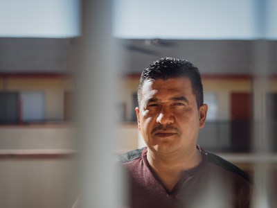 A person stands outdoors in partial shadow, framed by vertical lines in the foreground. The person has a calm expression and is looking forward. In the background, a building with windows and doors is softly out of focus under clear daylight.