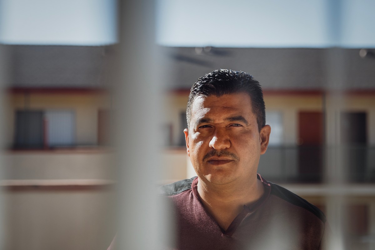 A person stands outdoors in partial shadow, framed by vertical lines in the foreground. The person has a calm expression and is looking forward. In the background, a building with windows and doors is softly out of focus under clear daylight.