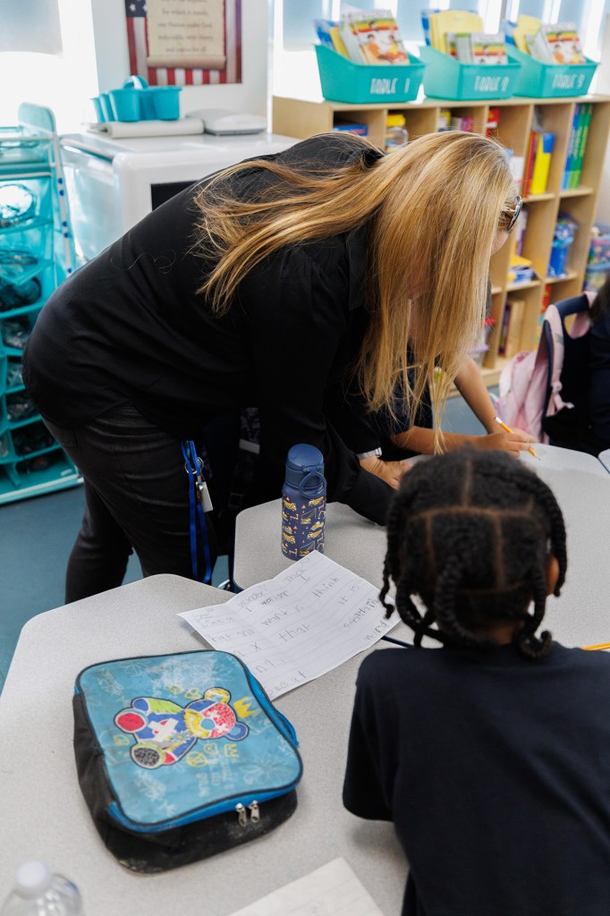 A teacher with blonde leaning down at a student's desk tp help them out. A worksheet, water bottle, a lunch bag and another student can be seen in the foreground.