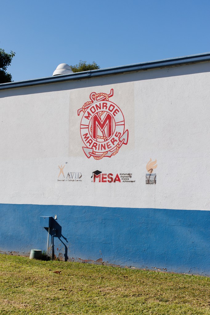 A red logo that reads "Monroe Mariners" among other smaller school-related logos is sen on the side of a white and blue colored building.