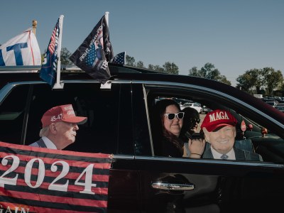 Two people inside a car look out the passenger window while one of them holds a cut out of President-elect Donald Trump wearing a hat that reads "MAGA". The outside of the car is decorated with more Trump paraphernalia including mini flags and a cut-out sticker of Trump pasted onto one of the side windows.