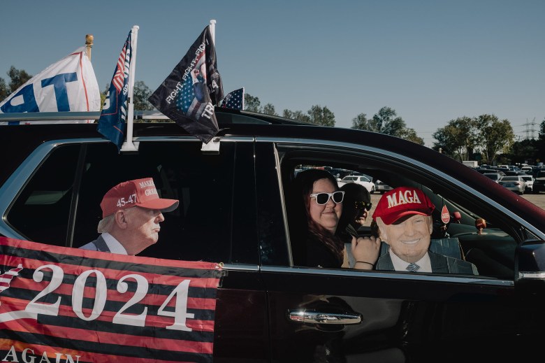 Two people inside a car look out the passenger window while one of them holds a cut out of President-elect Donald Trump wearing a hat that reads "MAGA". The outside of the car is decorated with more Trump paraphernalia including mini flags and a cut-out sticker of Trump pasted onto one of the side windows.