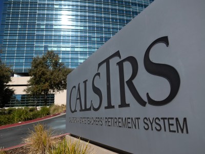 A sign for the California State Teachers' Retirement System (CalSTRS) stands prominently in the foreground, displaying the organization’s name in large, bold letters. Behind the sign, a modern glass building with reflective windows rises into the sky, partially shaded by nearby trees. Landscaping around the sign includes decorative grasses and shrubbery.