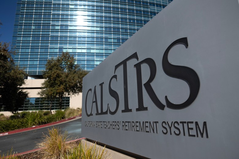 A sign for the California State Teachers' Retirement System (CalSTRS) stands prominently in the foreground, displaying the organization’s name in large, bold letters. Behind the sign, a modern glass building with reflective windows rises into the sky, partially shaded by nearby trees. Landscaping around the sign includes decorative grasses and shrubbery.