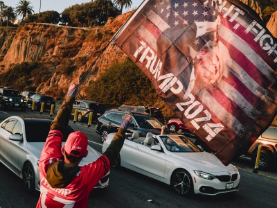 A person wearing a red 'Trump' hat and jersey holds a large flag that reads 'Trump 2024' with an image of Donald Trump over a U.S. flag backdrop. They face a line of cars, some with passengers who have their arms raised, as they drive along a coastal road bordered by rugged cliffs and tall palm trees, illuminated by warm sunlight.