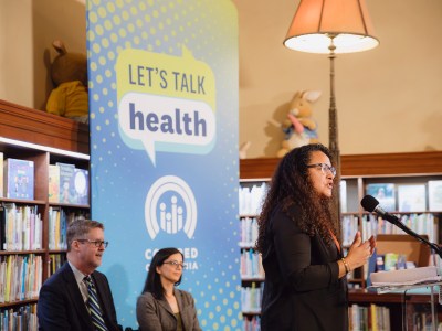 People on a small stage, with one person standing in front of a lectern, in front of an audience at a library surrounded by books and Covered California signage that reads "Let's talk health."