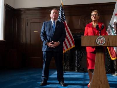 A person stands in the middle of a room with their hands folded as another person stands talking into a microphone at a podium.