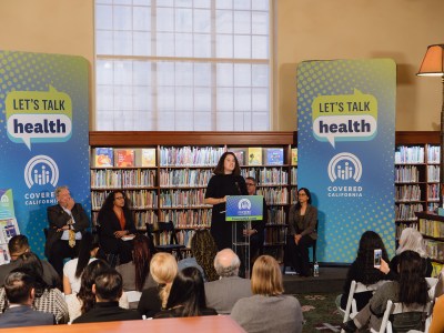 A wide view of people on a small stage, with one person standing in front of a lectern, in front of an audience at a library surrounded by books and Covered California signage that reads "Let's talk health."