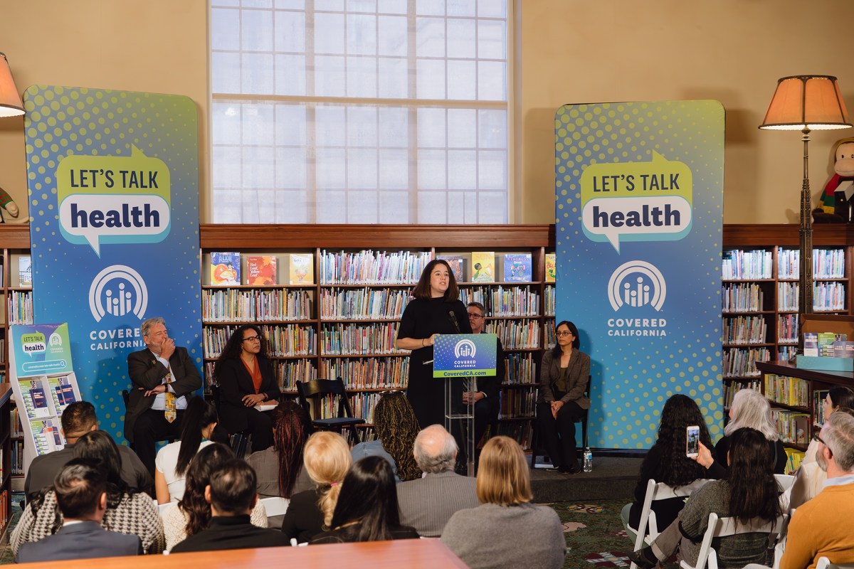 A wide view of people on a small stage, with one person standing in front of a lectern, in front of an audience at a library surrounded by books and Covered California signage that reads "Let's talk health."