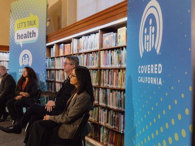 A close-up view of people on a small stage during an event at a library surrounded by books and Covered California signage that reads "Let's talk health."