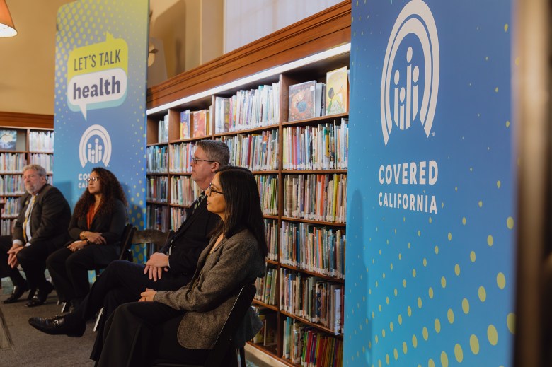 A close-up view of people on a small stage during an event at a library surrounded by books and Covered California signage that reads "Let's talk health."