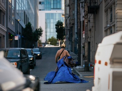 A person wearing a beige jacket and cap walks down a city street, pulling a large, crumpled blue tarp. The scene is framed by tall buildings, parked cars, and a modern glass structure in the background. The muted urban setting is illuminated by soft, natural light, highlighting the quiet and solitary moment.