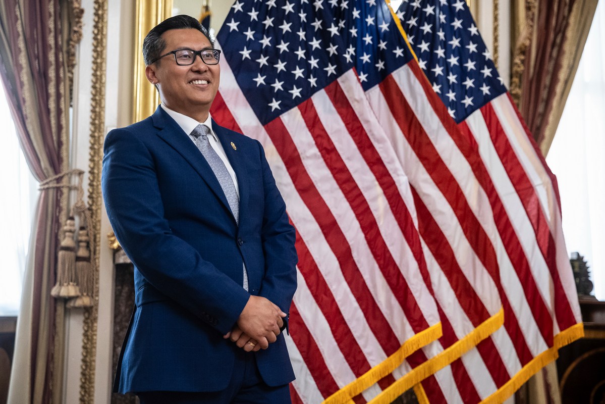 A person wearing glasses and dressed in a blue suit stands with his hands crossed in front of a row of American flags in a decorated office.