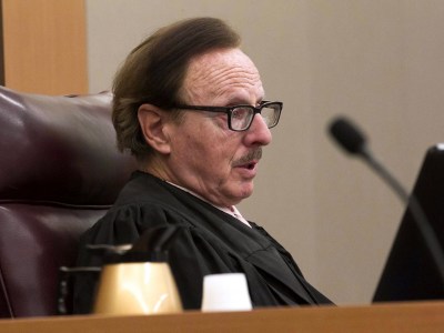 A person seated in a leather chair at a judicial bench, wearing a black robe and glasses. They appear to be engaged in court proceedings, with a microphone and a laptop visible on the desk in front of them. A light-colored wall and a portion of a wooden desk complete the courtroom setting.