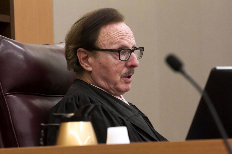 A person seated in a leather chair at a judicial bench, wearing a black robe and glasses. They appear to be engaged in court proceedings, with a microphone and a laptop visible on the desk in front of them. A light-colored wall and a portion of a wooden desk complete the courtroom setting.