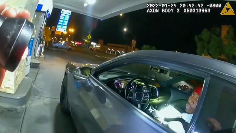 A nighttime scene captured from a body camera showing a car parked at a gas station. The vehicle's driver's side window is open, and the driver is looking toward the camera. The dashboard and steering wheel are visible inside the car. The gas station and surrounding area are illuminated, with signs and storefronts visible in the background.