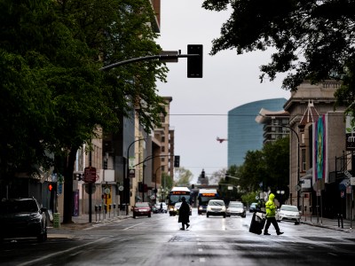 Two people are crossing the street in almost deserted downtown Sacramento, and cars are driving down the street.