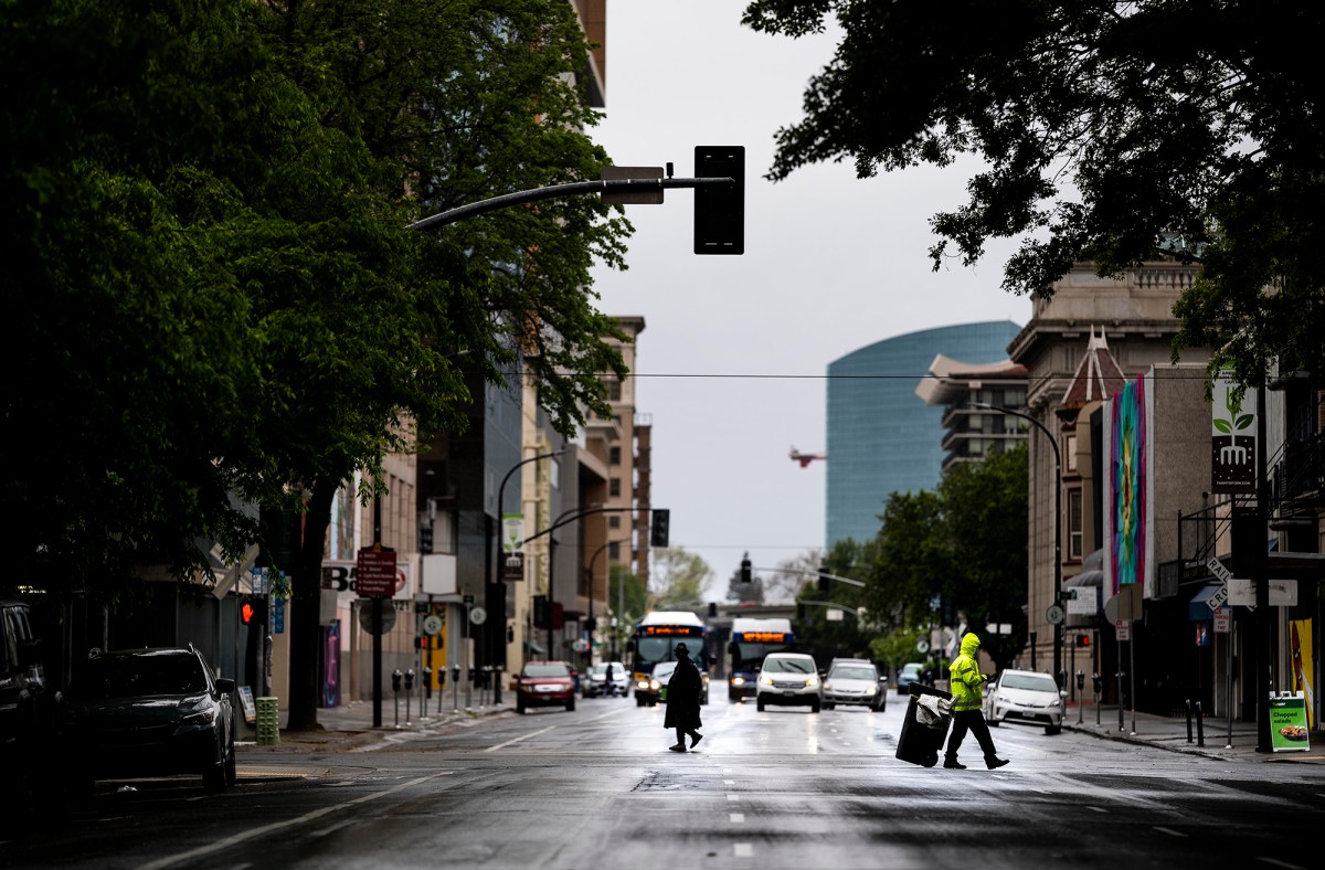 Two people are crossing the street in almost deserted downtown Sacramento, and cars are driving down the street.