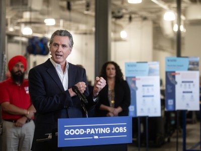 A person in a suit speaks at a podium labeled "GOOD-PAYING JOBS," gesturing with one hand. Behind them, two individuals stand near informational boards with text and graphics. The setting appears to be an indoor event space with industrial lighting.