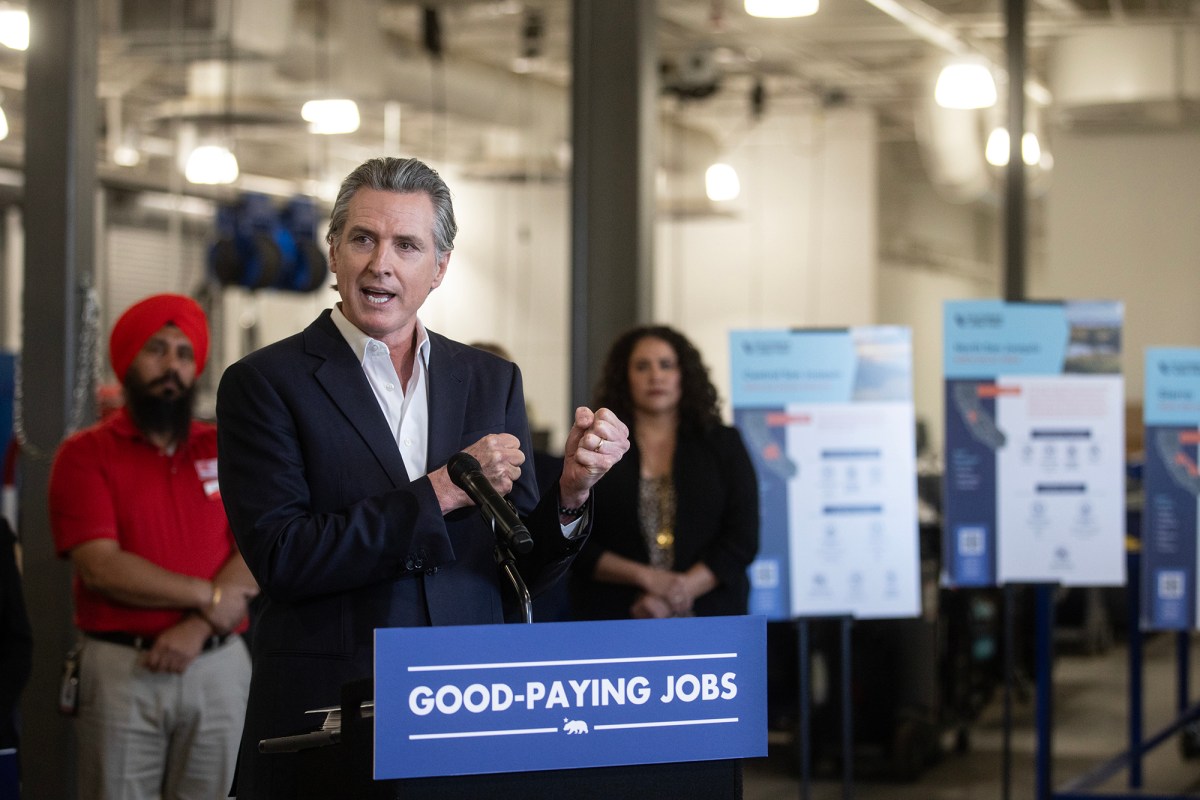 A person in a suit speaks at a podium labeled "GOOD-PAYING JOBS," gesturing with one hand. Behind them, two individuals stand near informational boards with text and graphics. The setting appears to be an indoor event space with industrial lighting.