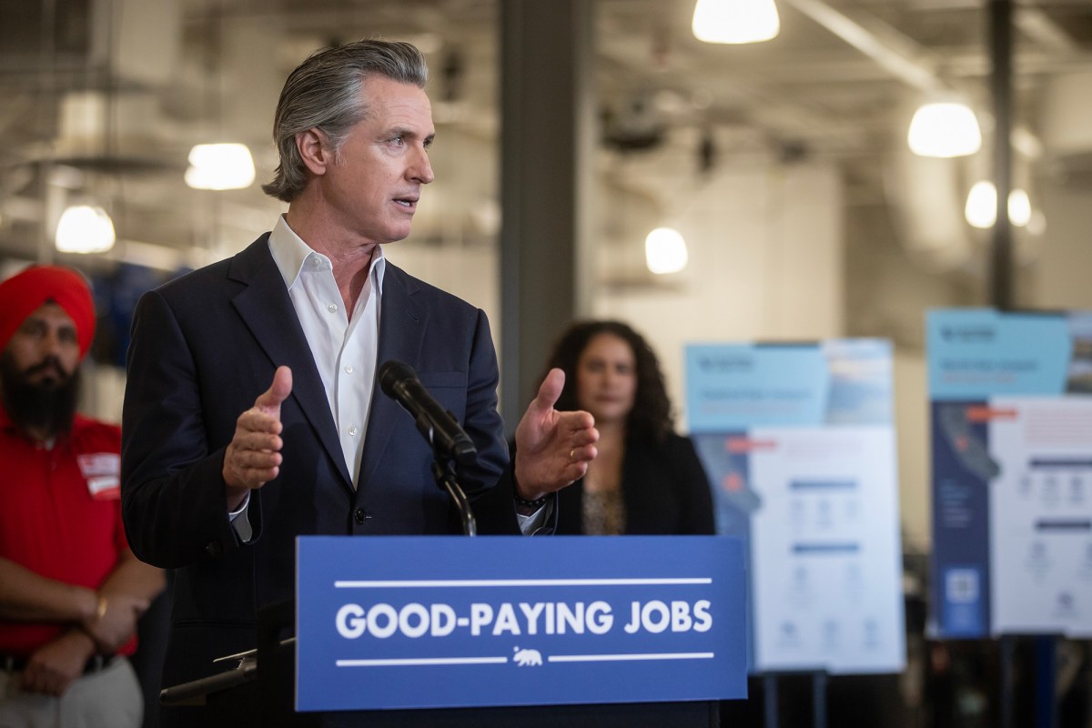 A person in a suit speaks at a podium labeled "GOOD-PAYING JOBS," gesturing with their hands. Behind them, two individuals stand near informational boards with text and graphics. The setting appears to be an indoor event space with industrial lighting.