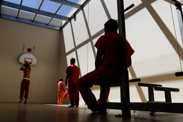 A person sits at a bench leaning against a pole as they watch a group of people play basketball in a room.