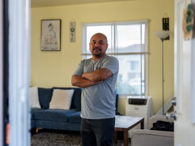 A view of a man wearing a gray shirt and black pants standing in the living room of his home. A frame Buddha art piece and decorations that read "love" and "home" can be seen in the background. Chanthon Bun stands in the living room of his home in San Leandro on Nov. 23, 2024. Bun was born in Cambodia during the genocide in the 1970s and grew up in a refugee camp in Thailand before moving to the United States, where he was convicted of a crime and at age 18. That was decades ago, but now he's anxious about President-elect Donald Trump's plan for mass immigrant deportation. Photo by Jungho Kim for CalMatters