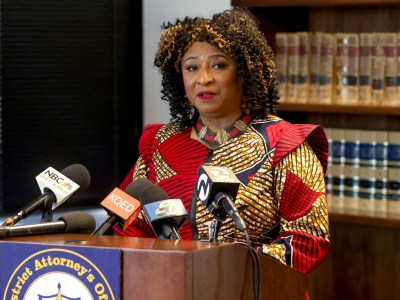 A person wearing a red and gold pattern dress stands behind a lectern with microphones from local media outlets during a press conference. A bookshelf with law books can be seen behind them.