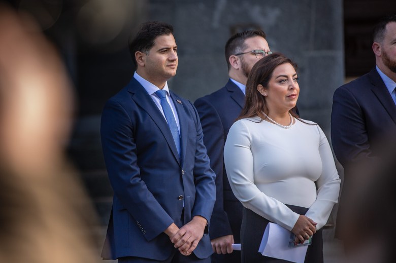 Two lawmakers, one wearing a blue suit and the other wearing a white and black dress, stand with their arms crossed in front of them, stand during a press conference at the state Capitol in Sacramento.