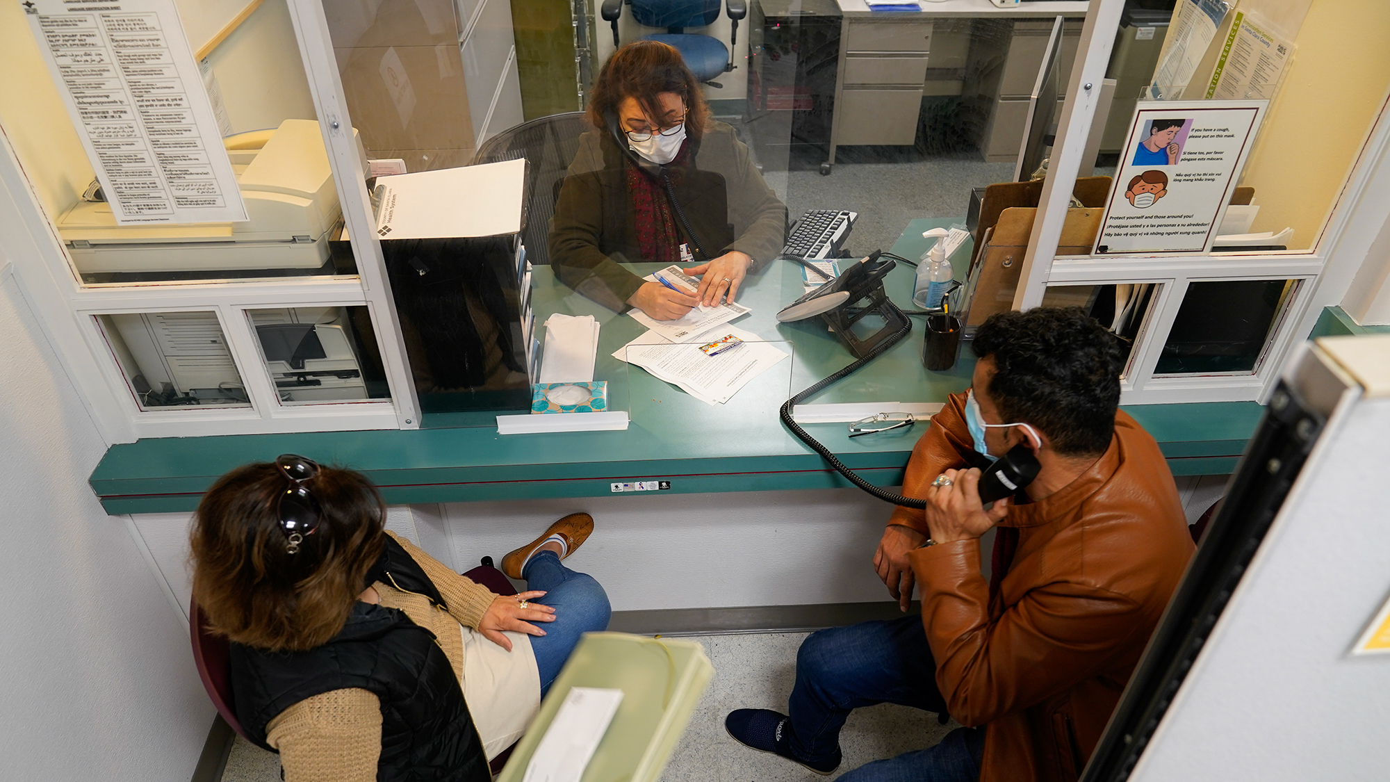 Two patients sit in front of a desk with a plexiglass where a receptionists writes something down on a piece of paper.